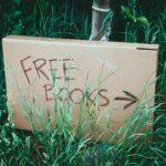 A cardboard sign with 'Free Books' handwritten, placed among green grass outdoors promoting book exchange.
