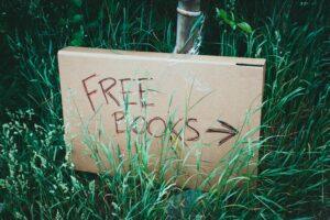 A cardboard sign with 'Free Books' handwritten, placed among green grass outdoors promoting book exchange.