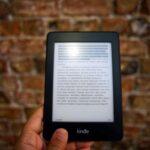 Close-up of a Kindle e-reader held by a person, showcasing a digital reading device against a textured brick wall backdrop.