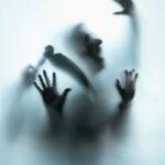 A mysterious silhouette with raised hands and a knife creates an eerie visual against frosted glass.